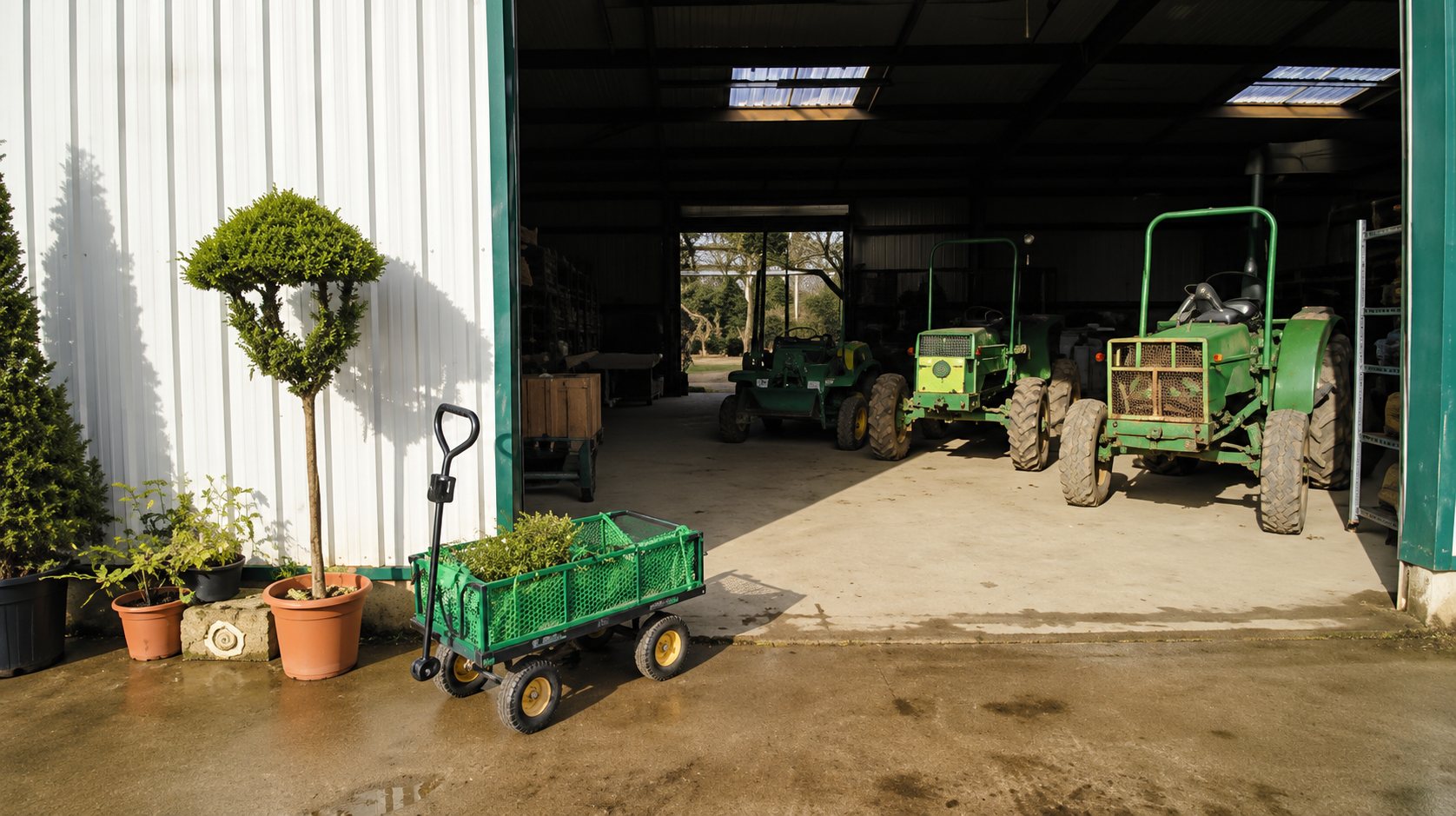 Devant l'ouverture d'un hangar, un chariot de jardin vert et des arbustes en pots sont posés près de tracteurs verts garés à l'intérieur.