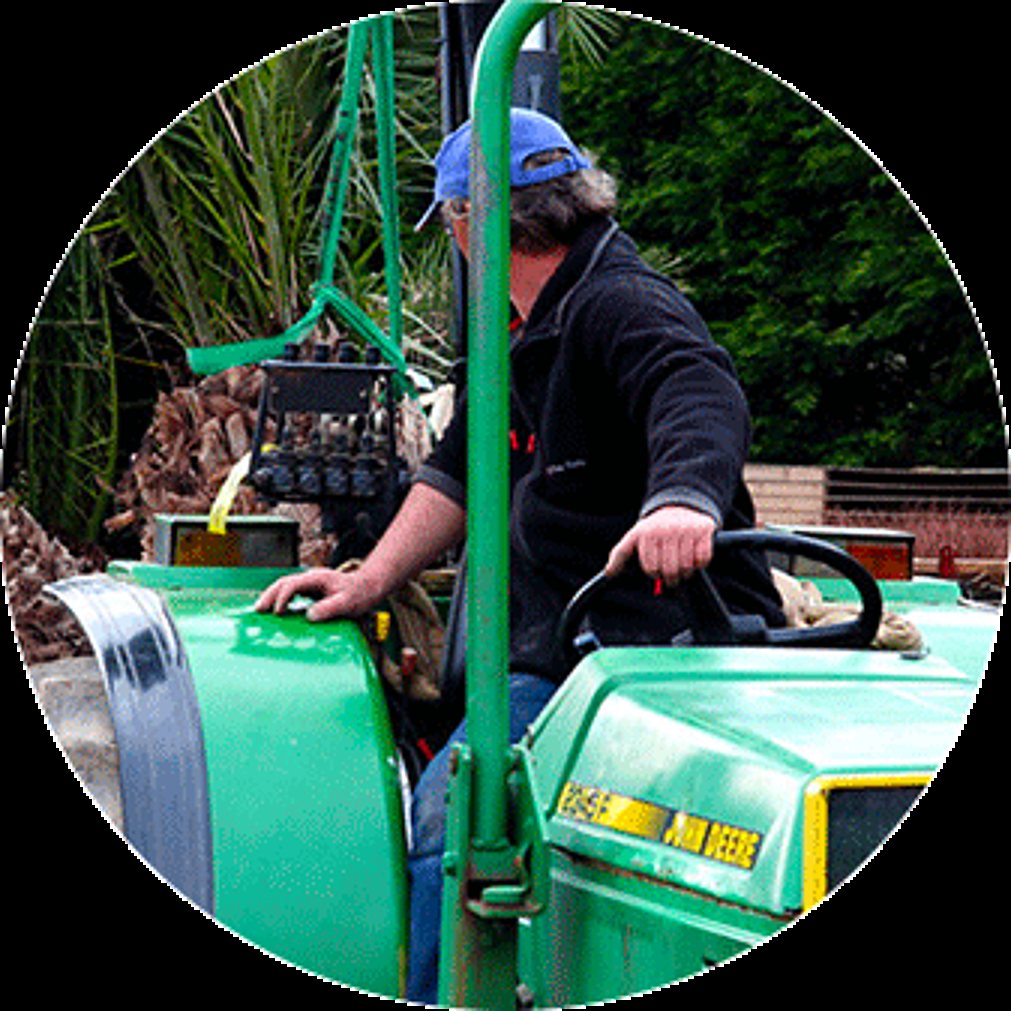 Un homme coiffé d'une casquette bleue conduit un tracteur John Deere vert, avec des palmiers et de la végétation en arrière-plan.