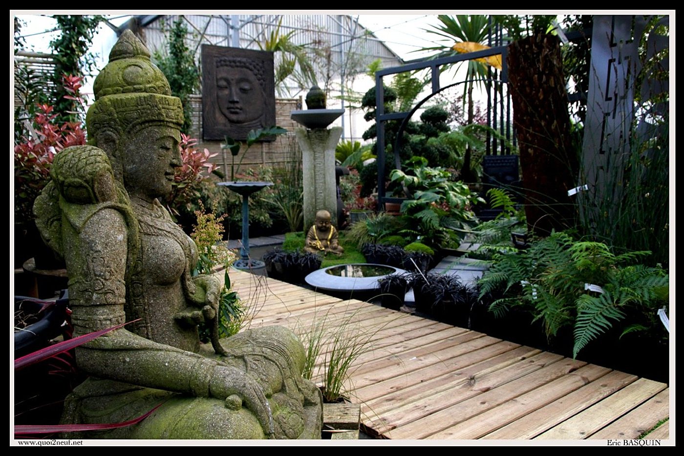Une statue de divinité en pierre couverte de mousse trône près d'une terrasse en bois, entourée de fougères, de plantes et d'un petit bouddha dans une serre aménagée en jardin zen.
