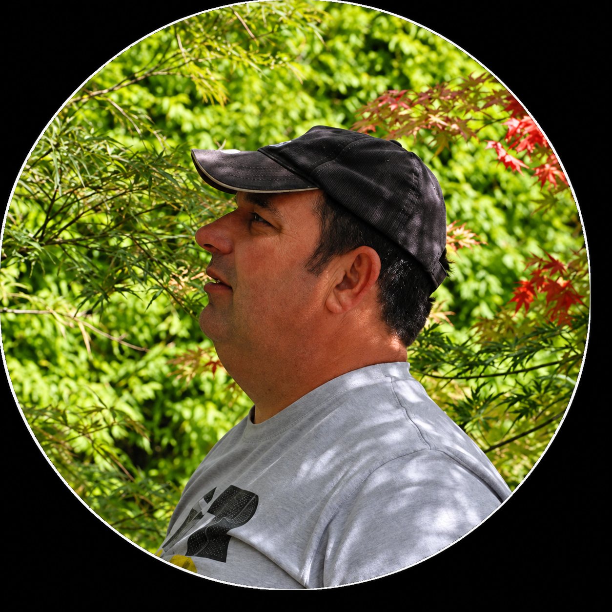 Un homme portant une casquette sombre et un t-shirt gris est photographié de profil devant un feuillage vert et rouge d'érable du Japon.