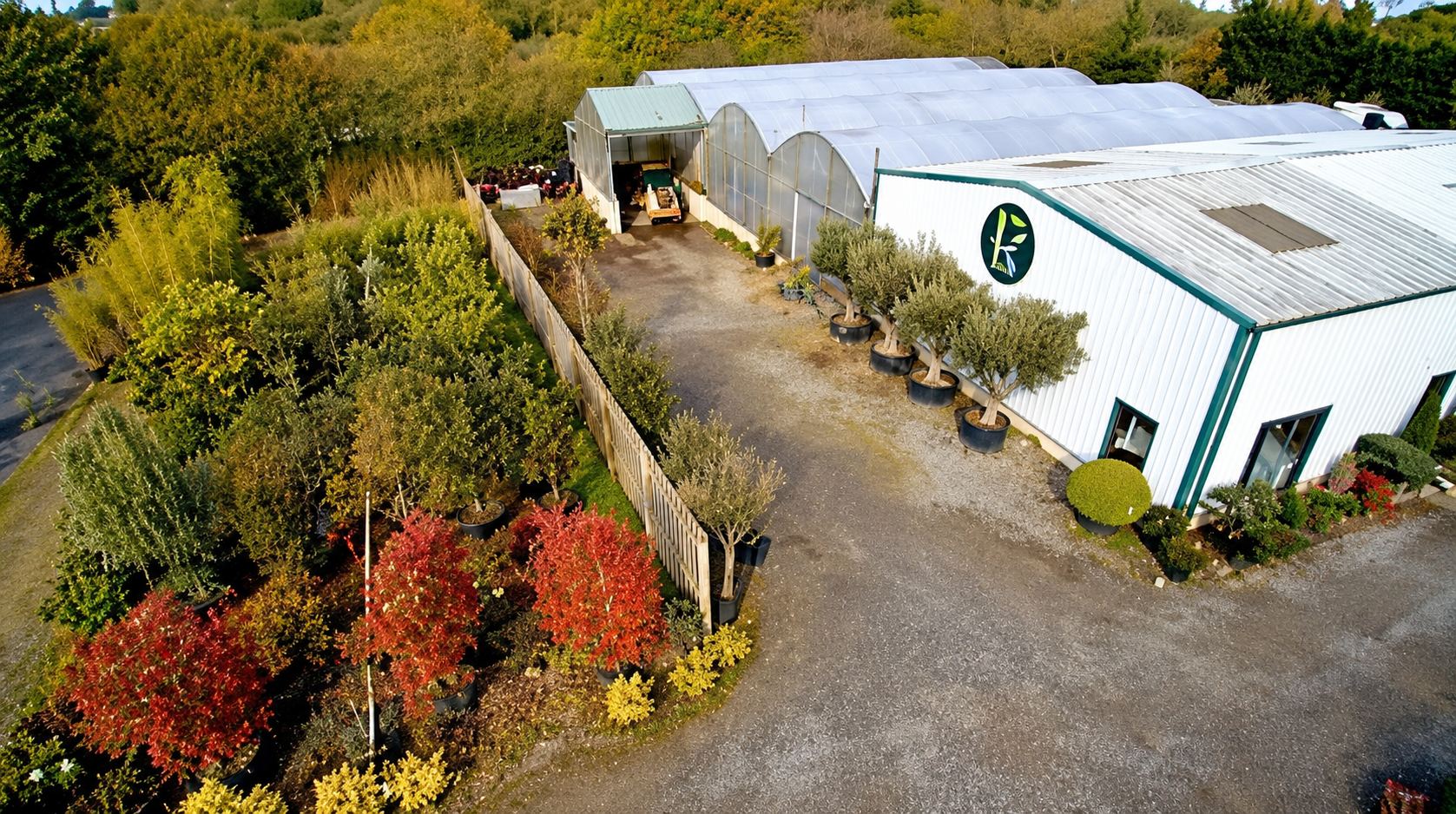 Vue aérienne d'une pépinière montrant des serres, un bâtiment blanc avec un logo circulaire, des arbres en pots et des arbustes aux feuillages colorés.