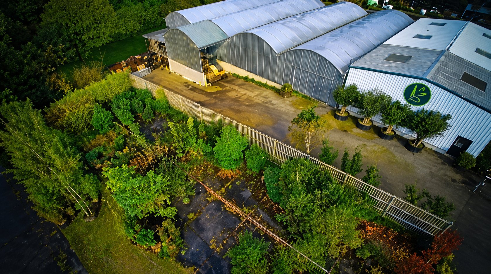 Vue aérienne prise au crépuscule montrant plusieurs serres en arc, un bâtiment avec un logo circulaire et un terrain planté de végétation dense au premier plan.